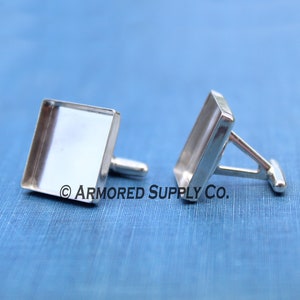 Puede incluir: Un par de gemelos de plata con inserciones de vidrio cuadradas. Los gemelos están sobre un fondo de tela azul. © ARMORED SUPPLY CO.