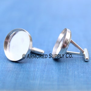 May include: Two silver-toned cufflinks with a round, flat surface and a post and toggle closure. The cufflinks are displayed on a blue textured background. The text "ARMORED SUPPLY CO." is visible.
