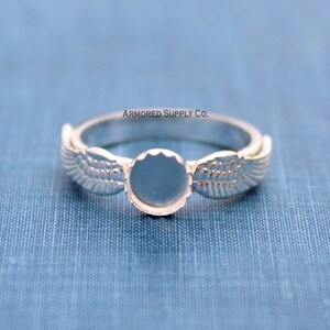 May include: Silver ring with a round, blank bezel and two wing designs on either side. The text "ARMORED SUPPLY CO." is printed above the ring.