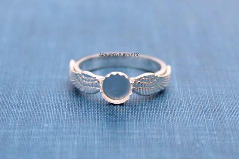 May include: Silver ring with a round, blank bezel and two wing designs on either side. The text "ARMORED SUPPLY CO." is printed above the ring.
