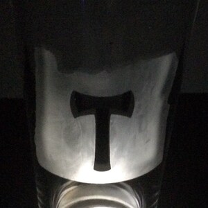 May include: A clear glass pint glass with a black silhouette of a hammer and a T inside. The glass is lit from below with a white light.