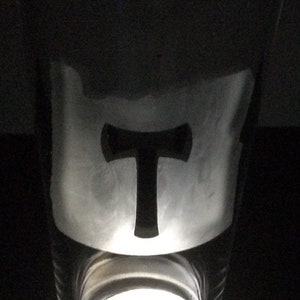 May include: A clear glass pint glass with a black silhouette of a hammer and a T etched on the side. The glass is illuminated from below by a white light.