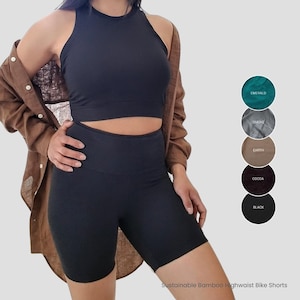 May include: Black high-waisted bike shorts made from sustainable bamboo. The shorts are shown on a model wearing a black tank top and a brown linen shirt. The image also shows a color chart with the following colors: Emerald, Smoke, Earth, Cocoa, and Black.