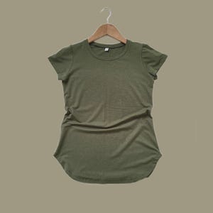 Peut inclure: T-shirt vert olive à col rond et manches courtes, suspendu à un cintre en bois. Le t-shirt a un ourlet incurvé et un aspect légèrement texturé. Le fond est de couleur neutre.