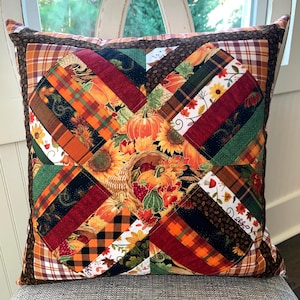 May include: A square throw pillow featuring a patchwork design with autumn-themed fabric patterns. The center displays a cornucopia with pumpkins, sunflowers, and fall leaves, surrounded by diagonal strips of plaid, checkered, and floral fabrics.