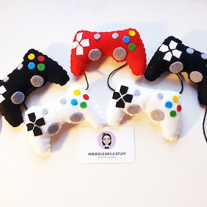 May include: Four felt game controllers in red, black, white, and black. The controllers are hanging from a string. The controllers have buttons and a directional pad. The controllers are handmade and are a fun and unique gift for gamers.