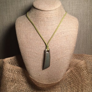 May include: A necklace with a rectangular pendant and a light green cord. The pendant is a muted green color with a textured surface. The cord is a light green suede-like material, and the necklace is displayed on a mannequin.