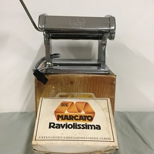 May include: A silver pasta maker with a crank handle, in its original box. The box is white with orange and yellow accents and says "Marcato Raviolissimma Ravioli Machine".