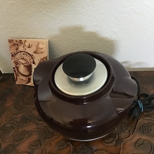 May include: A dark brown ceramic slow cooker with a black lid and a silver knob. The slow cooker is sitting on a wooden table with a carved design.