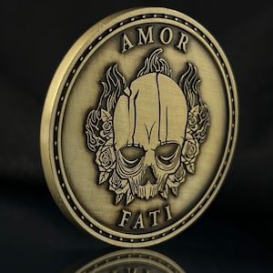 May include: A gold-toned metal coin with a skull design surrounded by roses and flames. The words "AMOR FATI" are inscribed on the coin.