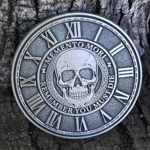 May include: A silver-toned round coin with a skull and crossbones design. The coin features a Roman numeral clock face with the text "Memento Mori - Remember You Must Die".