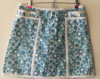 Peut inclure: Une mini-jupe bleue et blanche à motif circulaire. La jupe est ornée de bordures blanches et de deux poches avec boutons.