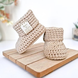 May include: A pair of beige crochet baby booties with a folded cuff. One bootie features a small rectangular tag with the text "Born In 2027" stitched onto the side. The booties are crafted from a textured yarn and have a rounded toe design.