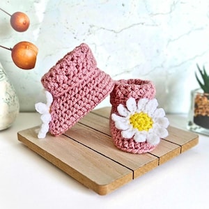 Crochet daisy baby booties, pictured in pale rose. With a white daisy that has a yellow centre. The booties are available in sizes newborn, 0-3 and 3-6 months.