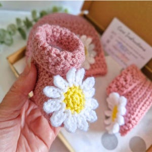 Crochet daisy baby booties, pictured in pale rose. With a white daisy that has a yellow centre. The booties are available in sizes newborn, 0-3 and 3-6 months.