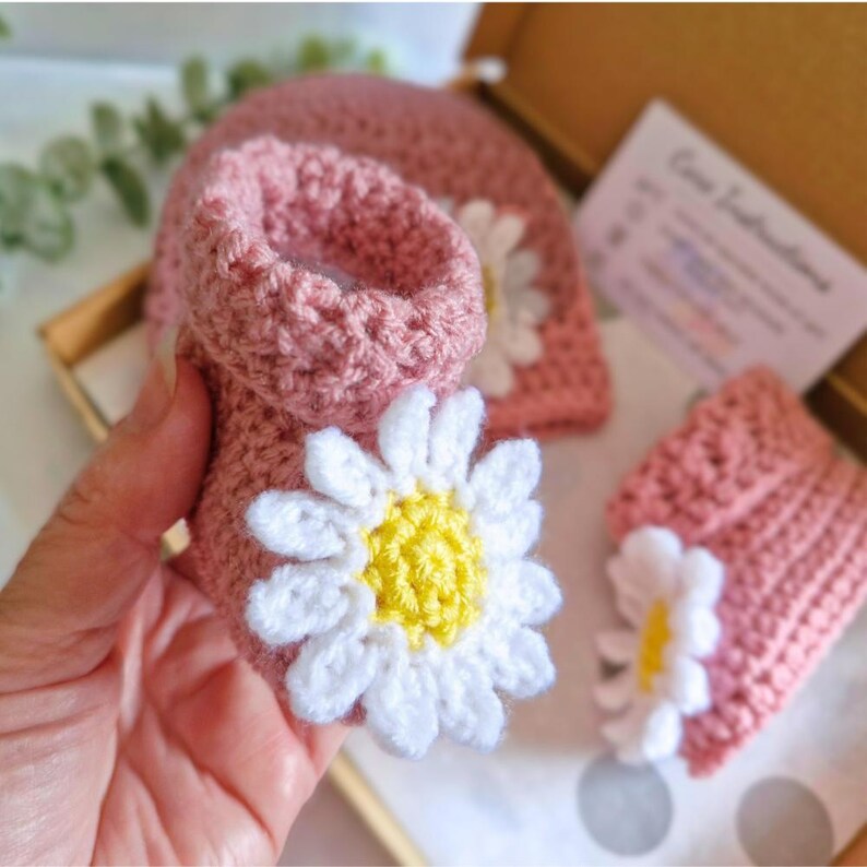 Crochet daisy baby booties, pictured in pale rose. With a white daisy that has a yellow centre. The booties are available in sizes newborn, 0-3 and 3-6 months.