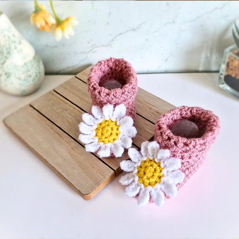 Crochet daisy baby booties, pictured in pale rose. With a white daisy that has a yellow centre. The booties are available in sizes newborn, 0-3 and 3-6 months.