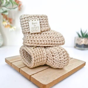 A pair of parchment crochet baby booties stacked on top of each other. The booties feature a small, rectangular, off-white tag attached to the side with the text Born In 2027 printed in a dark brown, simple sans-serif font.