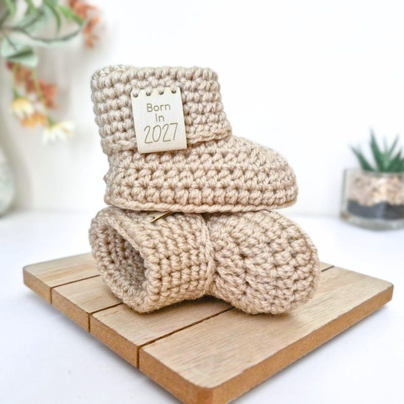 A pair of parchment crochet baby booties stacked on top of each other. The booties feature a small, rectangular, off-white tag attached to the side with the text Born In 2027 printed in a dark brown, simple sans-serif font.