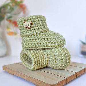 May include: A pair of light sage green crochet booties featuring a textured knit pattern and a small, circular wooden button with an engraved bear face design attached to the side of the upper cuff of the top bootie in the stack.