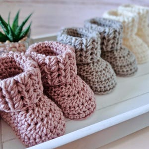 May include: A row of three pairs of handmade crochet baby booties arranged in a line. The first pair is dusty rose, the second is light gray, and the third pair is cream. Each pair features a thick, folded cuff and a textured, chunky knit design.