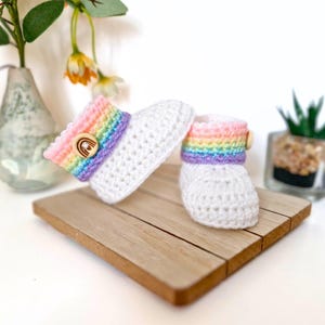 May include: A pair of white crocheted baby booties with rainbow trim and wooden buttons.