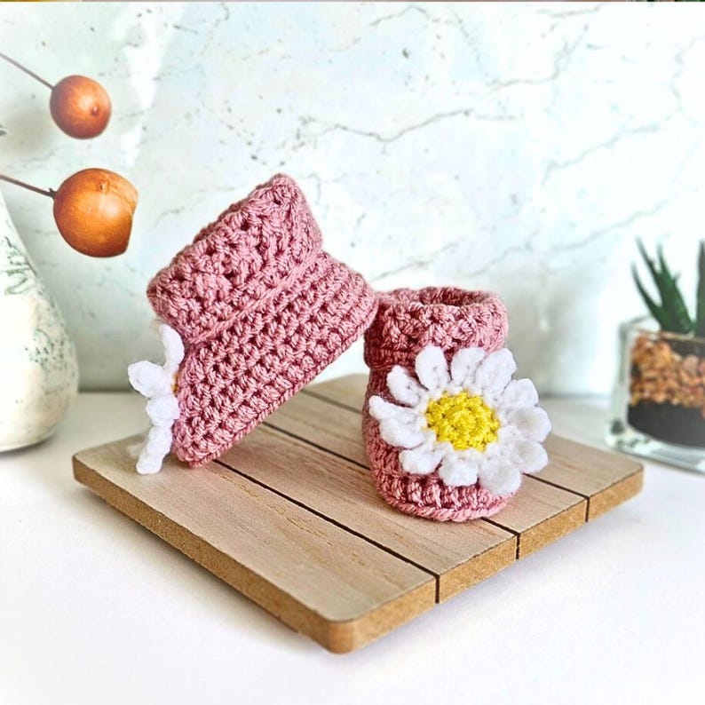 Crochet daisy baby booties, pictured in pale rose. With a white daisy that has a yellow centre. The booties are available in sizes newborn, 0-3 and 3-6 months.