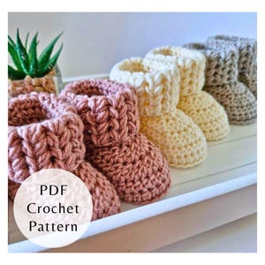 May include: A collection of handmade crochet baby booties in various colors, including pink, cream, and gray. The booties are arranged in a row, showcasing their textured stitch patterns. The image also includes a small succulent plant in a decorative pot. The text "PDF Crochet Pattern" is visible.