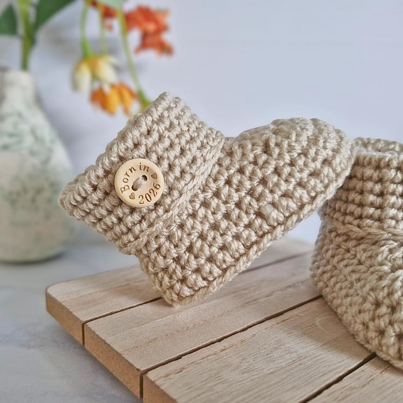 side view of a pair of crocheted baby booties in a light brown colour. The booties have a fold-over cuff and a button detail.