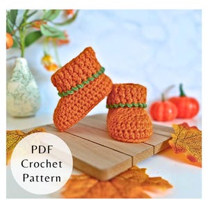 Puede incluir: Un par de botines de bebé de ganchillo naranja con ribete verde. Los botines se muestran sobre una superficie de madera con un fondo borroso de calabazas y follaje otoñal. El texto "PDF Crochet Pattern" es visible.