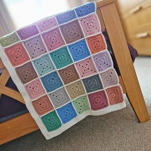May include: A colourful, handmade crochet blanket with a patchwork design. The blanket features squares in pastel shades of pink, blue, green, and brown, with a white border. It is draped over a wooden bed frame.