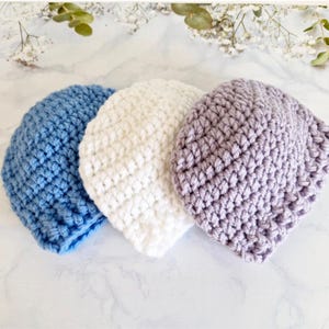 May include: Three crocheted beanies in shades of blue, white, and lavender are arranged on a marble surface. The hats are handmade with a textured stitch pattern. The beanies are a soft, cozy accessory.