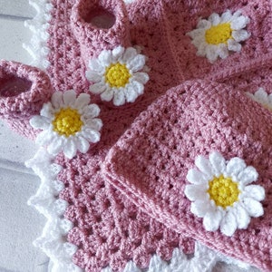 A pink crocheted baby hat with a white daisy flower design. The hat is paired with a pink crocheted baby bootie with a white daisy flower design.