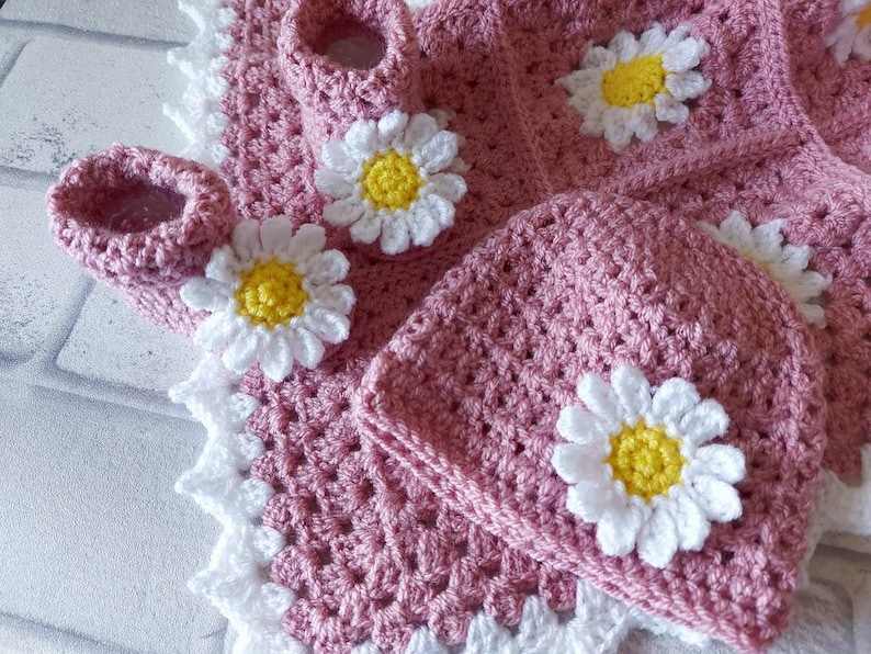 A pink crocheted baby hat with a white daisy flower design. The hat is paired with a pink crocheted baby bootie with a white daisy flower design.