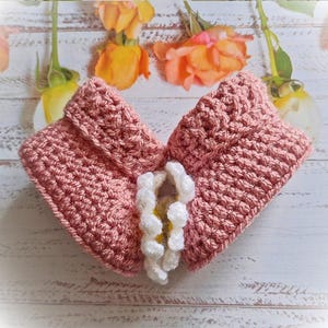 Side view of a pair of crochet daisy baby booties, pictured in pale rose. With a white daisy that has a yellow centre. The booties are available in sizes newborn, 0-3 and 3-6 months.