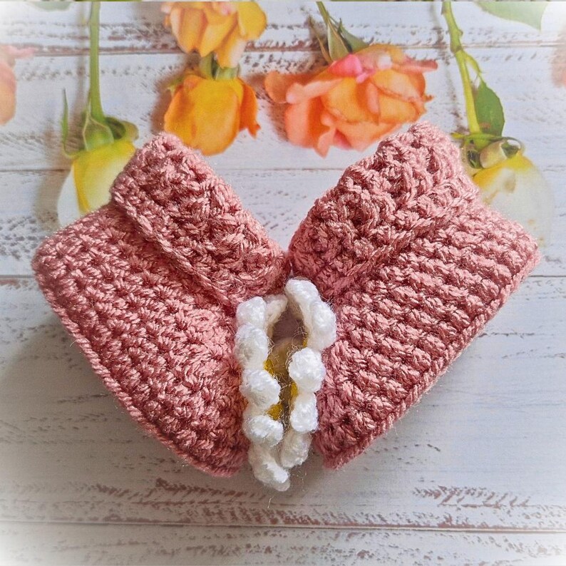 Side view of a pair of crochet daisy baby booties, pictured in pale rose. With a white daisy that has a yellow centre. The booties are available in sizes newborn, 0-3 and 3-6 months.