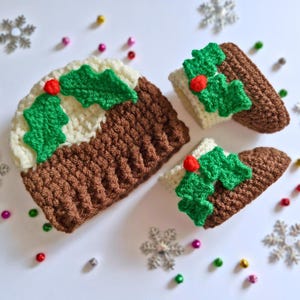 May include: Crocheted Christmas pudding decorations. The brown pudding has a white top with green holly leaves and red berries. Two smaller pudding decorations are also present. The decorations are on a white surface with snowflake and colorful bead accents.