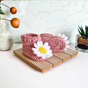 Crochet daisy baby booties, pictured in pale rose. With a white daisy that has a yellow centre. The booties are available in sizes newborn, 0-3 and 3-6 months.