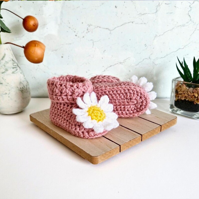 Crochet daisy baby booties, pictured in pale rose. With a white daisy that has a yellow centre. The booties are available in sizes newborn, 0-3 and 3-6 months.