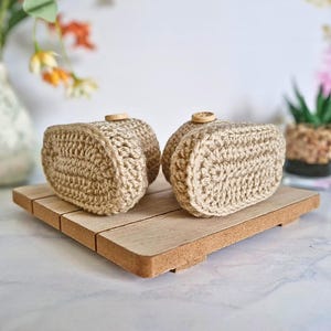 Close up view of the soles of a pair of beige crocheted baby booties with buttons on the top. Booties are on a wooden tray with flowers in the background.