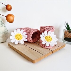 Crochet daisy baby booties, pictured in pale rose. With a white daisy that has a yellow centre. The booties are available in sizes newborn, 0-3 and 3-6 months.