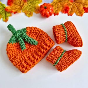 May include: A crochet set consisting of an orange hat with a green stem and leaf detail on top, accompanied by two matching orange booties featuring a green horizontal stripe around the cuff, all crafted with a textured yarn stitch pattern.