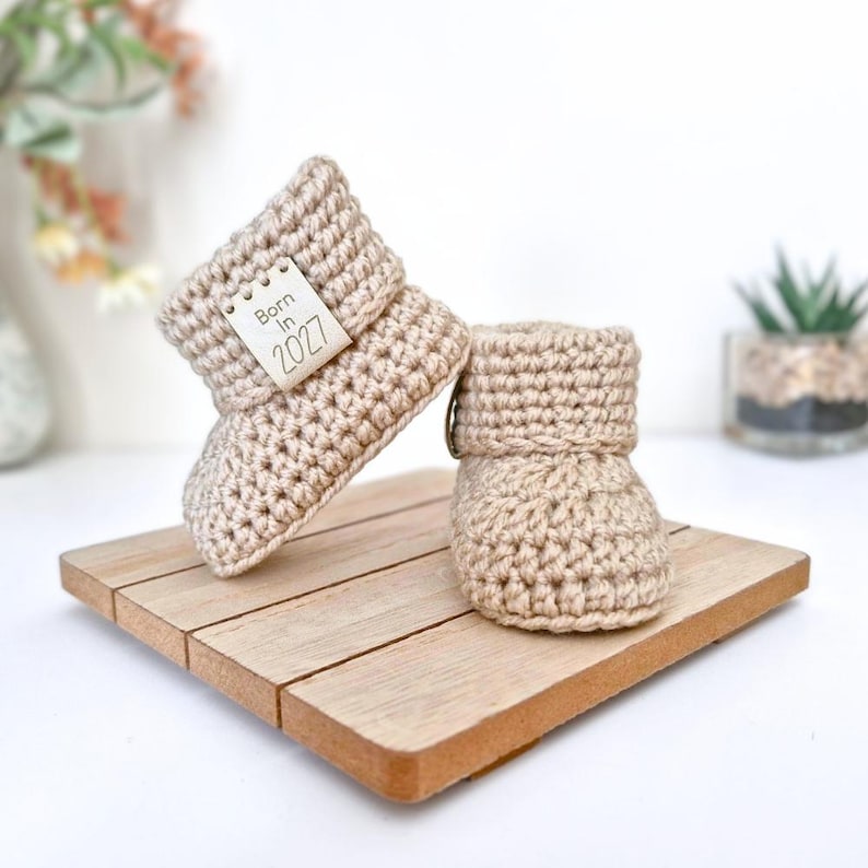 Handmade crochet baby booties with a neat folded cuff, finished with Born in 2027 faux leather tags, sizes newborn, 0-3 and 3-6 months and multiple colour choices - cute baby shower gift