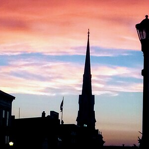 Peut inclure: Silhouette d'un haut clocher d'église sur fond de ciel de coucher de soleil coloré. Le ciel est un mélange de rose, d'orange, de bleu et de violet. Un lampadaire se trouve au premier plan.