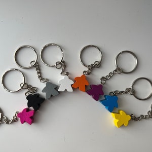 May include: A set of eight keychains featuring colourful wooden figures. The figures are shaped like people and are attached to silver key rings. The colours of the figures are white, orange, purple, blue, yellow, pink, black, and grey.