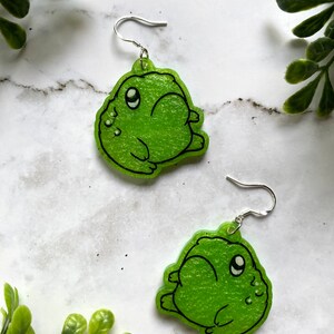May include: A pair of green frog dangle earrings with silver hooks. The frogs have black eyes and a textured surface.