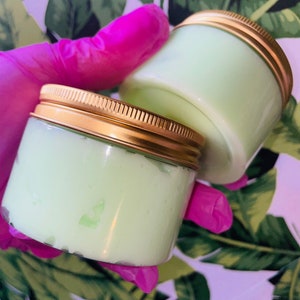 Puede incluir: Dos pequeños frascos de vidrio con tapas doradas llenos de una crema verde claro. Los frascos se sostienen con un guante de látex rosa.