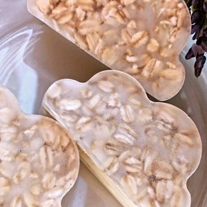 May include: Three cloud-shaped soaps with a light brown oatmeal embedded in the clear soap. The soaps are on a white surface.