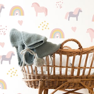 May include: A woven wicker baby bassinet with a grey knitted blanket and pom-poms. The background features a repeating pattern of pink horses, rainbows, hearts, and yellow dots on a cream wall. The bassinet is made of woven brown wicker.