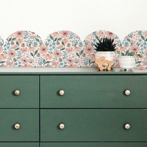 May include: A dark green six-drawer dresser with gold knobs, set against a white wall. Above the dresser is a floral wallpaper border with pink and blue flowers. A woven pendant light hangs from the ceiling, and a toy horse stands to the left.